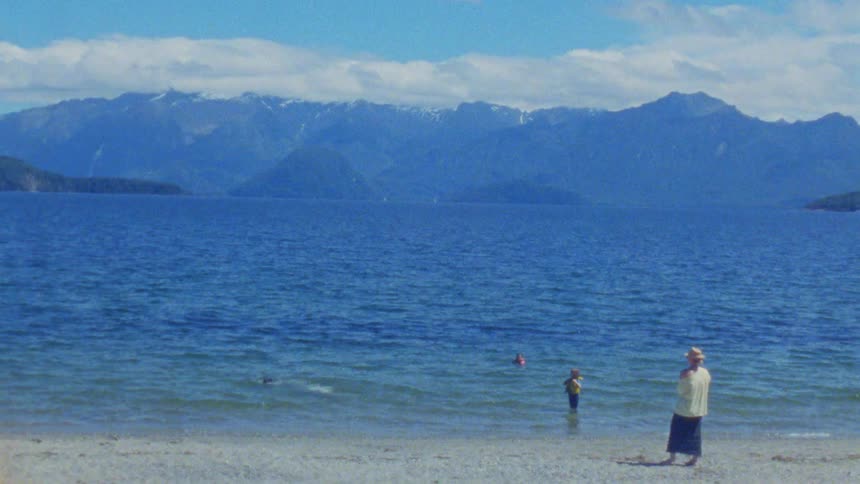 Person watching children and a dog playing in the lake with mountains in the background