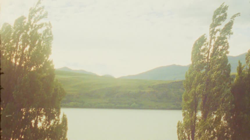 Lake surrounded by tall trees with green hills and mountains in the background under partly cloudy sky