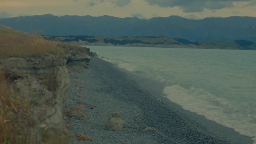 A view of a rocky beach shows waves crashing on the shore with a steep cliff on the left and mountains in the distance under a cloudy sky during the day