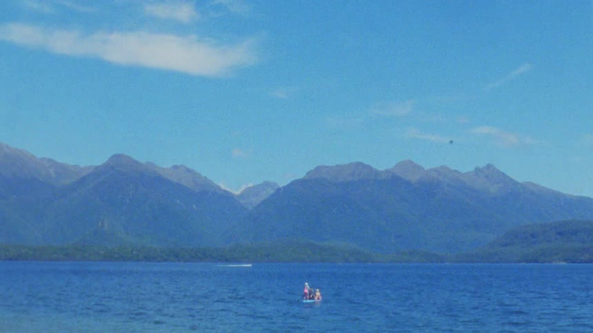 Person in small boat at center of lake with mountains in background under blue sky