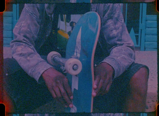 Vintage film footage shows the hands of a person sitting down while holding a blue skateboard with white wheels