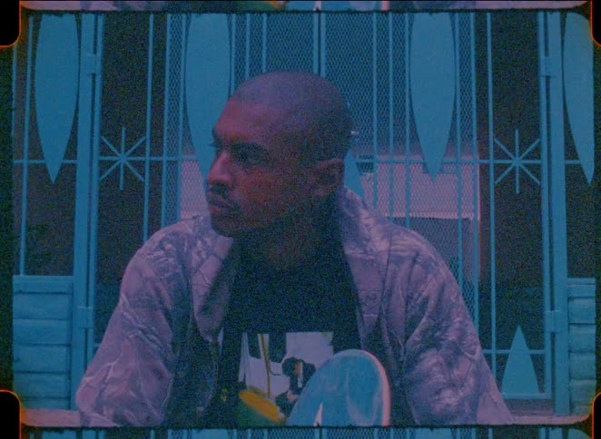 A young man sitting outdoors with a skateboard captured in a vintage film aesthetic with heavy grain and cool blue tones he appears contemplative as he looks offcamera resting his hands on the board while seated in front of a blue gate