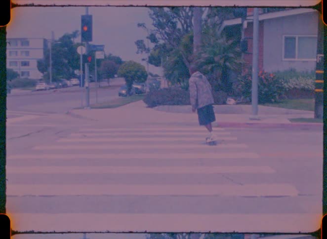 This video features a person on a skateboard riding across a residential street intersection it is shot with a vintage grainy film aesthetic that gives the scene a nostalgic or retro feel