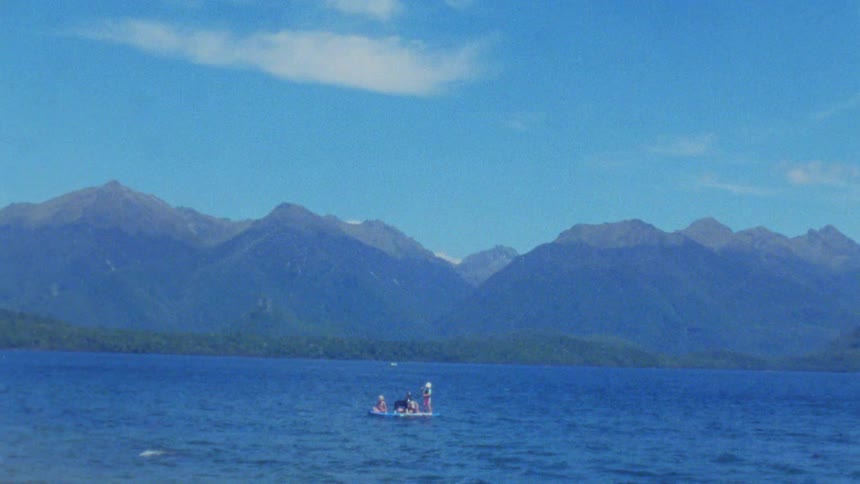 A group of people sits on a small boat in the middle of a large blue lake surrounded by dense green forests and high mountain peaks under a clear sky