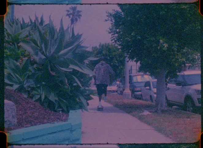 A young black male wearing a jacket riding a skateboard on the sidewalk with cars parked along the road and trees and grass lining the street in a suburban community at midday