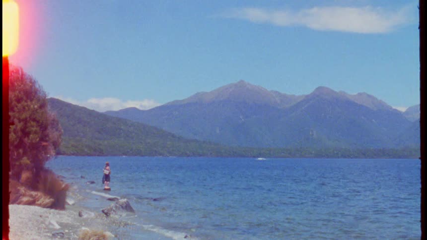 A person standing on a rock by a lake as the wind creates ripples on the water with a shoreline lined with trees and a mountain range on the opposite side at midday