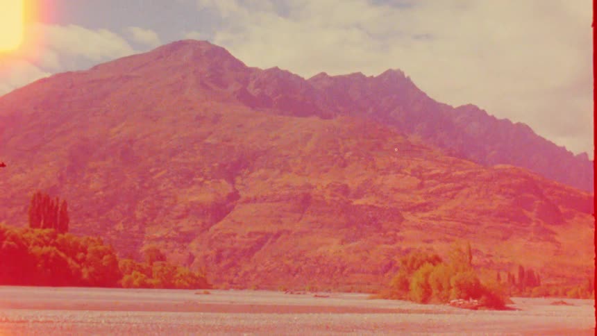 Vintage film footage shows a yellow jet boat speeding across a shallow river with a rocky shore in front of green trees and large mountains under a bright sky