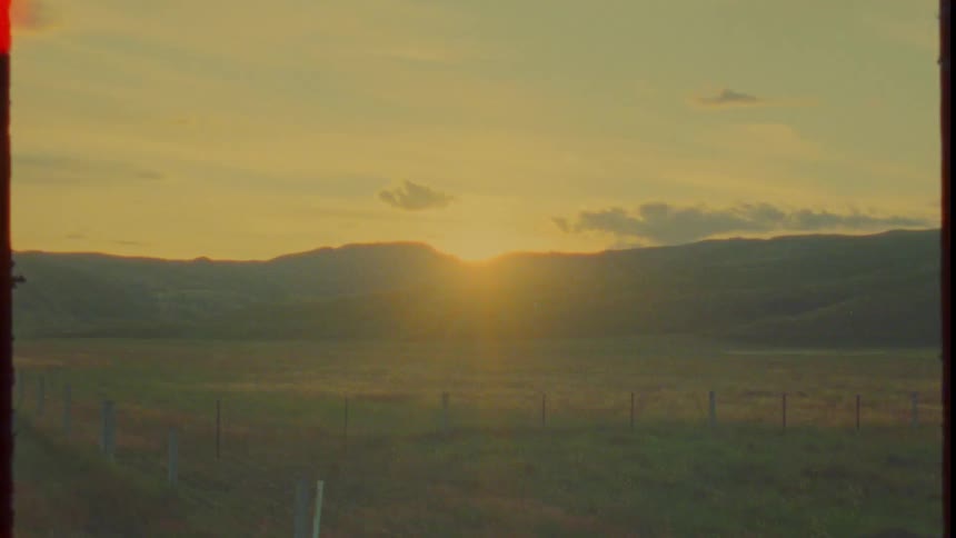The sun hangs low over a wide field and distant hills as redorange film burn and light leaks creep in from the edges washing the scene into a textured amber frame that ends like exposed reel footage