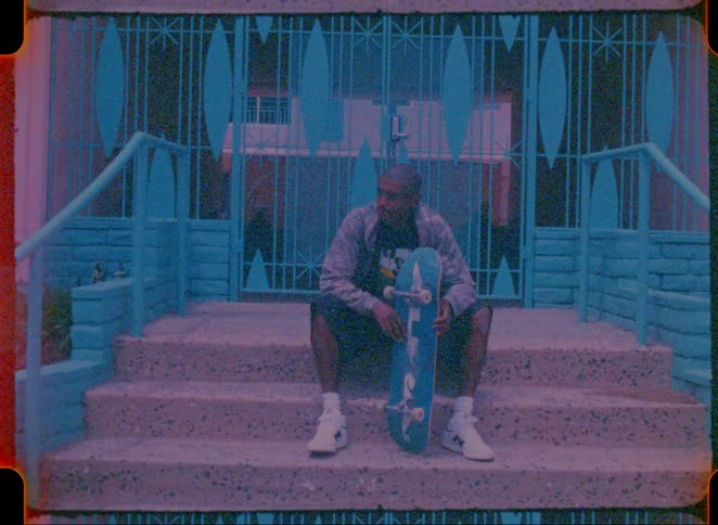 A man sits on concrete steps holding a skateboard in front of a blue metal gate during the day