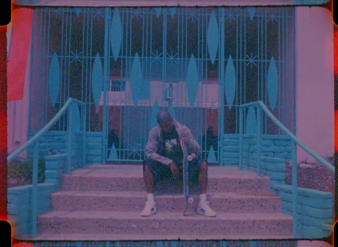 A man wearing a grey jacket and shorts sits on concrete steps while holding a skateboard in front of a blue decorative gate with light leaks on the film frame
