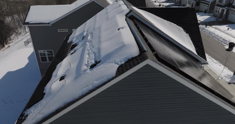 The drone footage reveals a striking contrast between the thick heavy snow blanketing the shingled roof and the sleek clear surface of the adjacent solar panels as the camera pans it captures the moment a massive shelf of snow loses its grip and slide