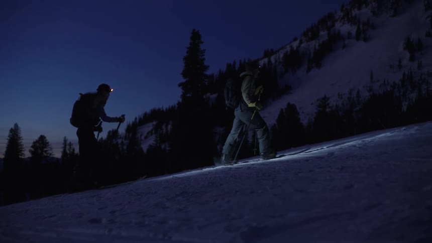 Two people ski on a snowy mountain at night with headlamps
