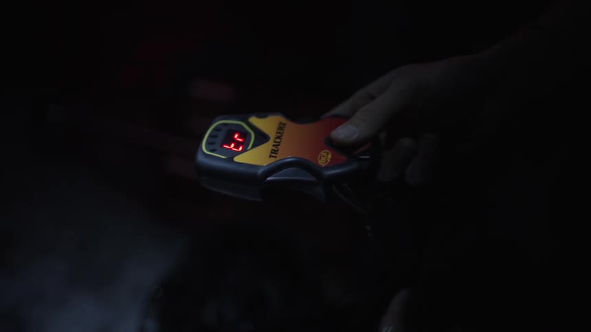 Aman holds a yellow and black tracker device in their hands during a dark night while pressing buttons and viewing a red led display screen before placing the electronic tool inside of a black bag