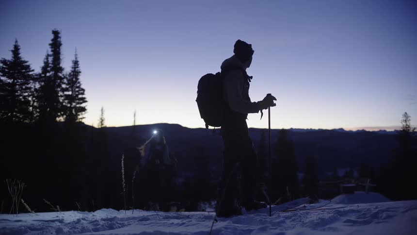 Two individuals with backpacks and poles walking in snowy landscape at dawn with pine trees and mountains in the background