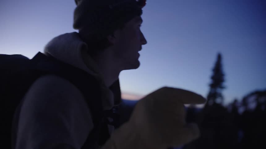 A male skier wearing a headlamp and backpack uses skins on his skis to climb a snow covered mountain during the sunrise