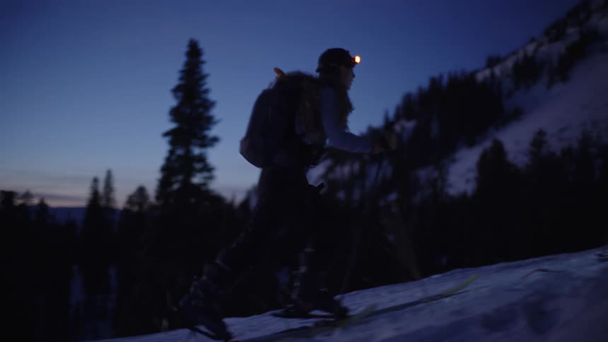 Two individuals climbing snowy slope at night with skis and poles illuminated by flashlight