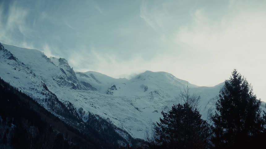 The shot shows a vast mountain range covered in snow under a pale blue sky where light clouds drift over sharp peaks while dark evergreen trees frame the bottom right corner