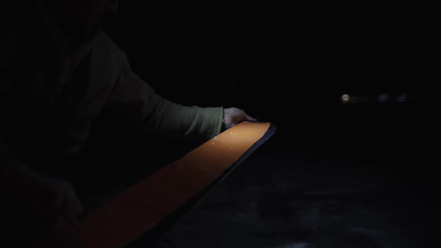 A man wearing a beanie and a headlamp applies orange climbing skins to a pair of skis in a dark outdoor setting at night