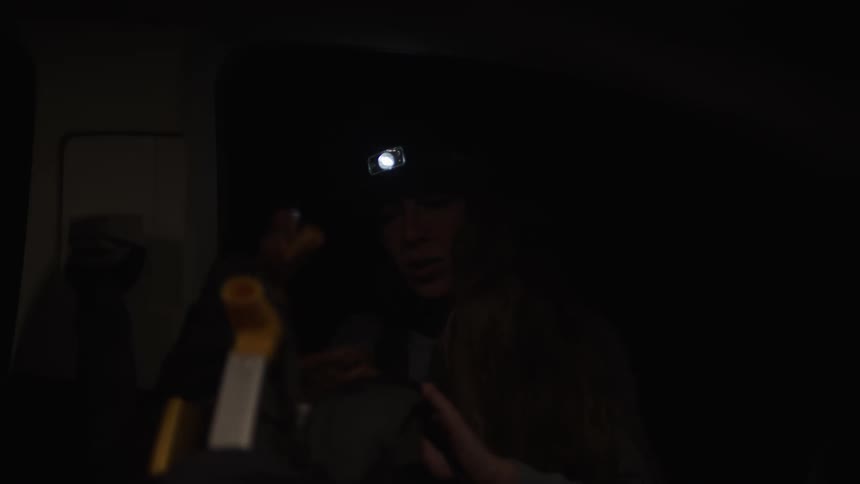 A woman wearing a headlamp sits in the dark interior of a vehicle while she organizes gear and packs a large backpack before a night excursion