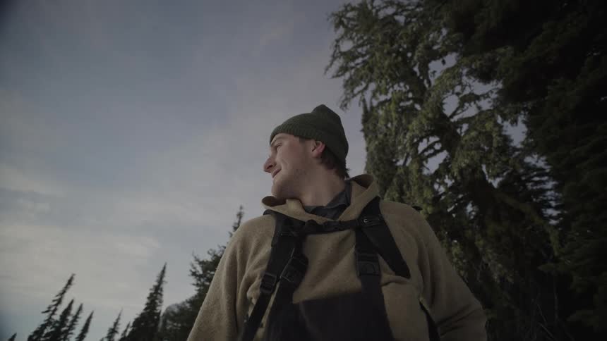 A man wearing a green beanie and a tan fleece jacket with a backpack stands in a snowy forest looking around at tall evergreen trees under a pale sky