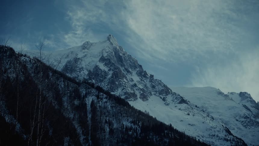 A snow covered mountain peak rises against a blue sky with thin white clouds while dark evergreen trees cover the lower slopes in a cold winter atmosphere