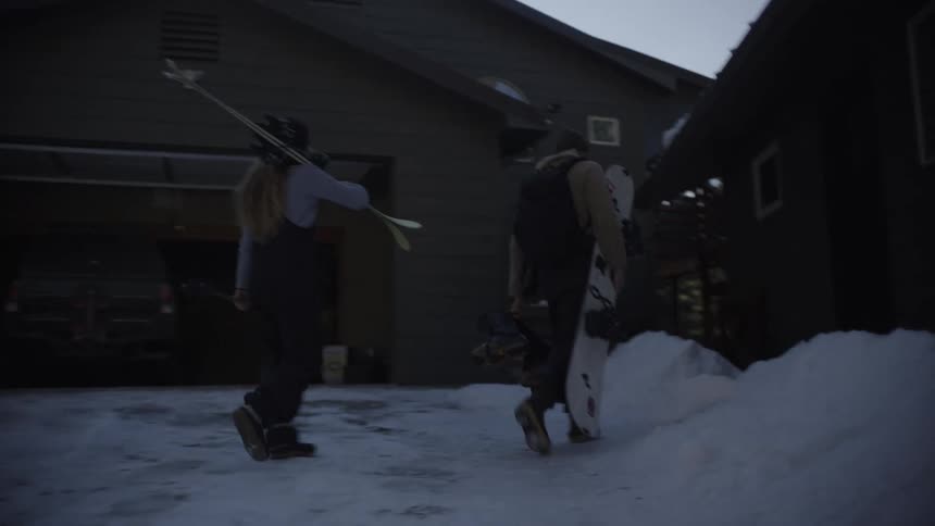 Two people carrying skis and a snowboard walk through deep snow toward a house on a sunny day