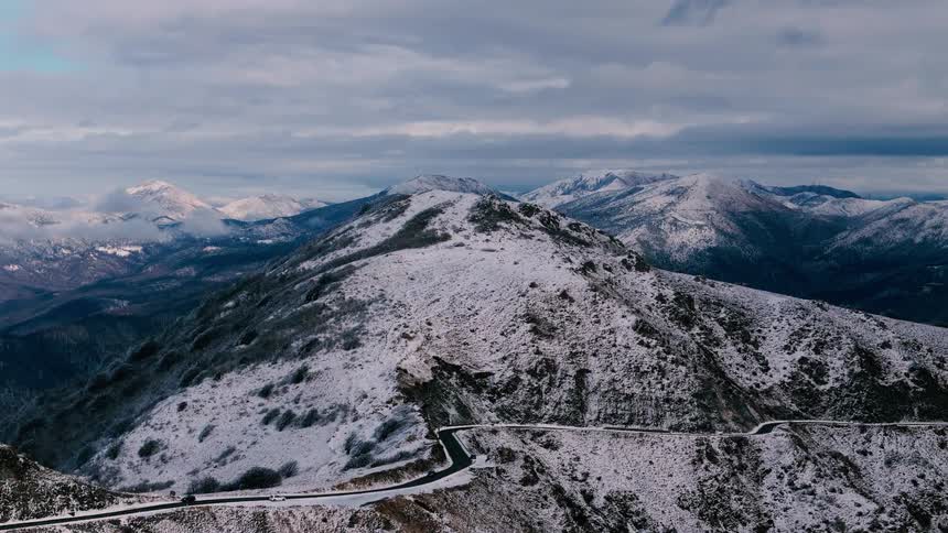As the camera glides over a rugged mountain landscape a winding asphalt road snakes across the snowdusted ridges leading the eye toward a series of majestic whitecapped peaks under a soft overcast sky