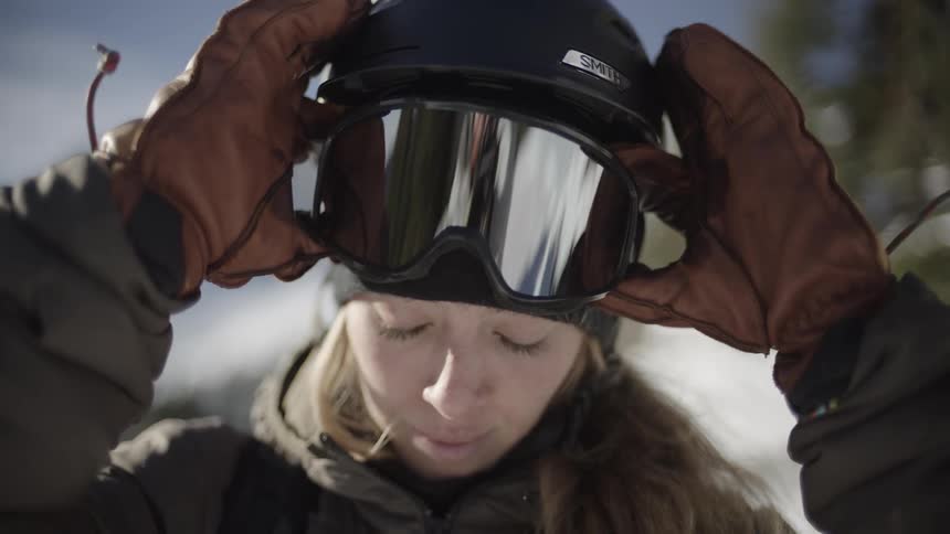 A woman wearing a black helmet and goggles over a warm jacket looks into the camera before pulling the goggles down over her eyes in a snowy mountain setting on a bright day