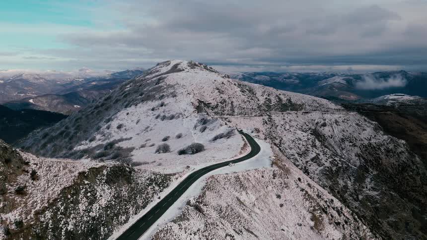 A view from above of a winding road along a mountain ridge covered in patches of snow under a grey and cloudy sky