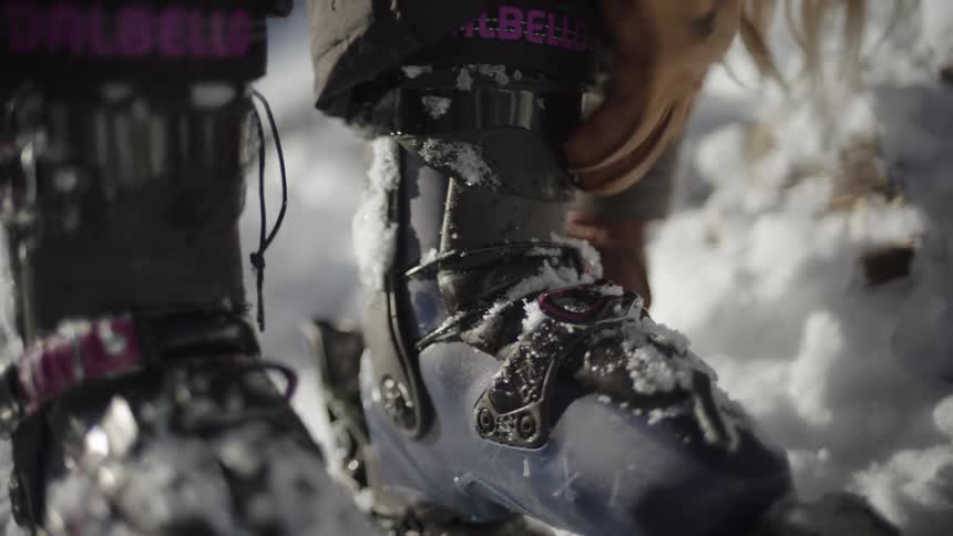 A close view of a person wearing tan leather gloves as they tighten the straps and buckles of their blue ski boots while standing in deep snow on a sunny day