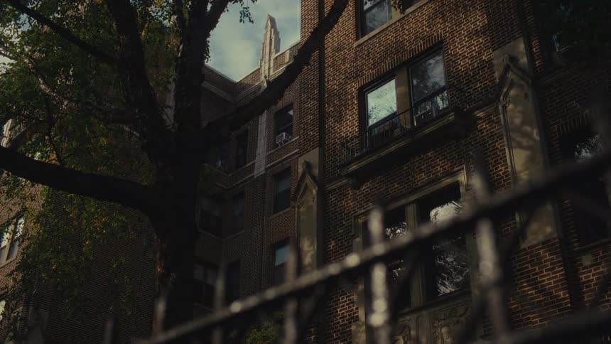 A view through a black metal fence of a large brick apartment building with many windows and a tall green tree in the foreground