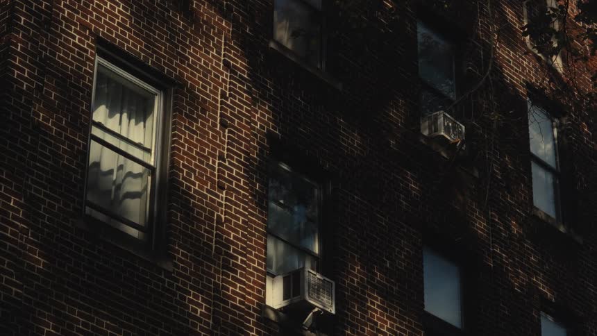 Golden sunlight and the shifting shadows of tree branches dance across the textured brick facade of an apartment building illuminating windows and air conditioning units in the quiet afternoon