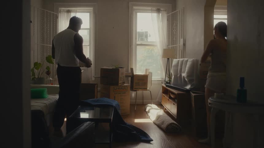 A man in a white tank top and a woman in a grey tank top and white shorts pack belongings into cardboard boxes in a bright apartment with large windows