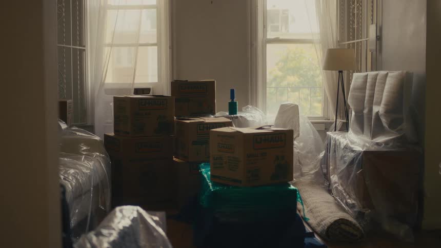 Moving boxes and furniture covered with plastic in a room lit by windows
