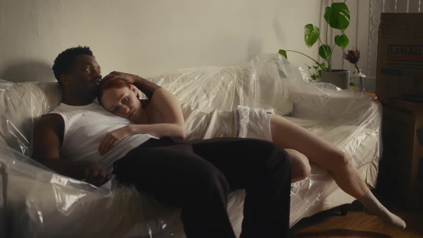 A man and a woman rest together on a sofa covered in protective plastic while taking a break from packing cardboard boxes in their apartment during the day