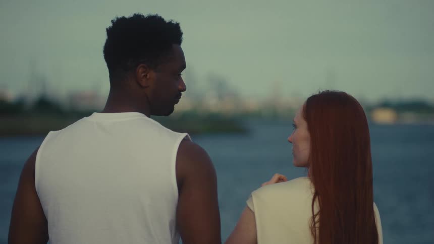 A man in a white tanktop and a woman with long red hair stand by a river at dusk and they are viewed from behind with a blurred city skyline across the water in the background until they get off the screen