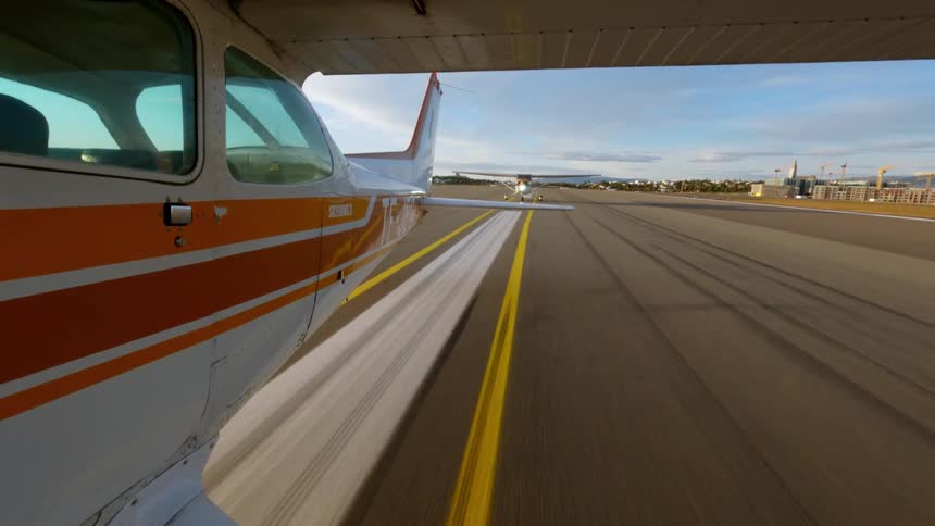 Viewed from the side of a small aircraft the plane speeds down a sunlit runway following bright yellow markings as another small plane waits in the distance near the airport terminal