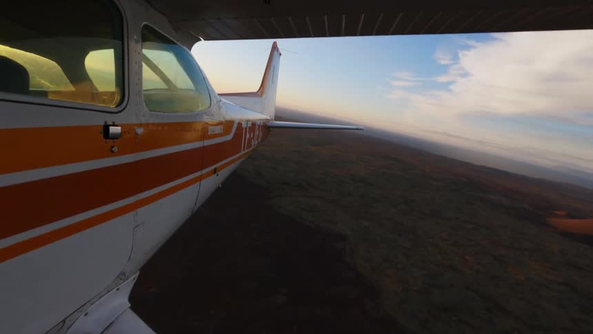 A view from a small white and orange airplane shows the wing and fuselage as it flies over a vast brown landscape toward a bright setting sun