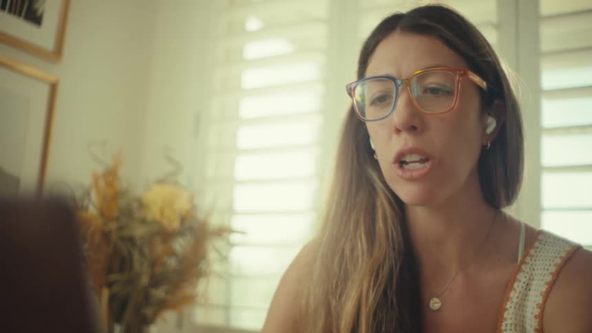 A woman with colorful glasses and earbuds talks during a video call in a bright room with window shutters and dried flowers in the background