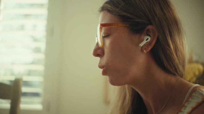A woman in amber glasses and earbuds sits at a dining table by a bright window speaking calmly toward someone offscreen as her expression shifts between serious and faintly amused