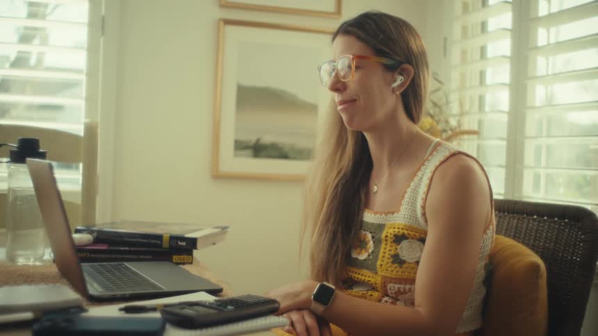 A woman wearing glasses and wireless earbuds sits at a table with a laptop and stacked books smiling slightly as sunlight streams through nearby windows