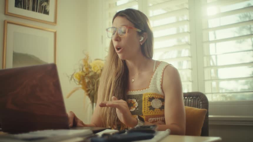 A woman wearing yellow glasses and a crochet top sits at a desk while speaking and gesturing toward her laptopthe room is brightly lit by natural light coming through window shutters creating a warm atmosphere as she appears to be on a video call