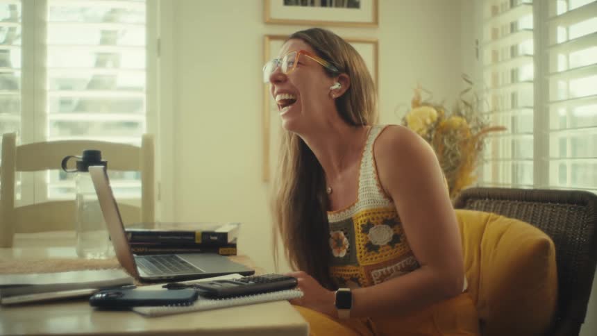 A woman wearing glasses and a crochet top laughs while sitting at a table with a laptop and books during the day