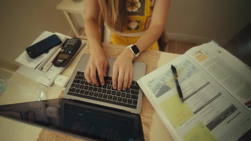 A woman types on a silver laptop at a wooden desk scattered with a calculator an open textbook and sticky notes