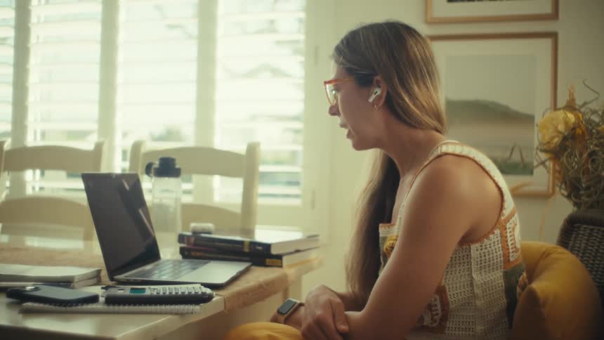 A young woman wearing bright glasses and earbuds sits at a dining table actively engaged in a video call on her laptopthe scene is set in a sunlit room with white shutters where she gestures with her hands while speaking to the person on the screen