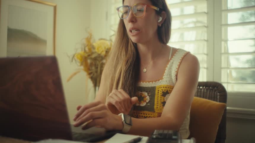 A woman wearing colorful glasses and wireless earbuds talks on her laptop at a bright table with papers nearby