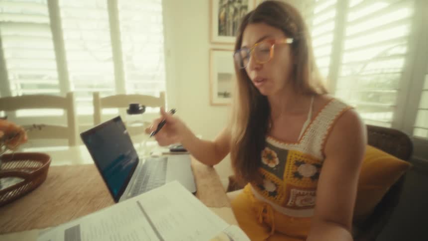 A woman wearing orange glasses and a crochet top turns the page of a study planner and writes with a pen at a wooden table while working near a bright window with white shutters