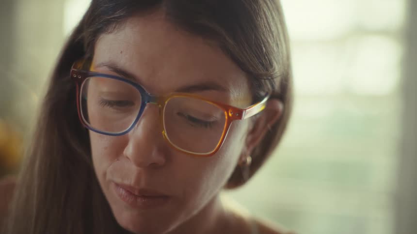 A woman wearing eyeglasses is sitting at a desk in a sunlit room typing ona calculator and writing in a notebook