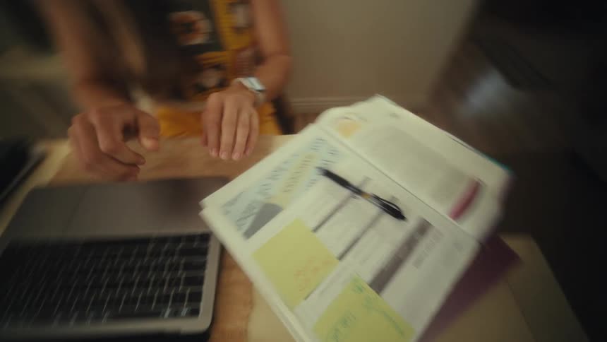 A young woman sitting behind a desk uses a pen to write on a document then turns to her computer and begins typing in an office at midday