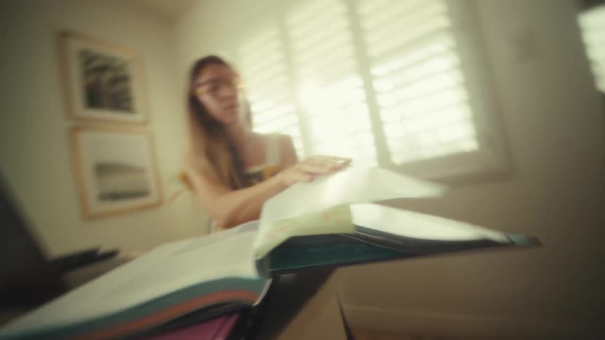 A view from a low angle shows a woman sitting at a table and flipping through the pages of a large open book during the day